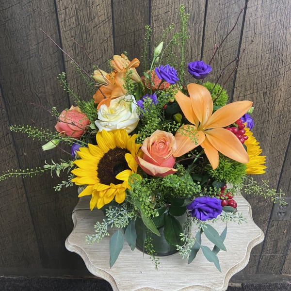 Mixed bouquet with sunflowers, orange lilies, roses, and purple blooms in a glass vase