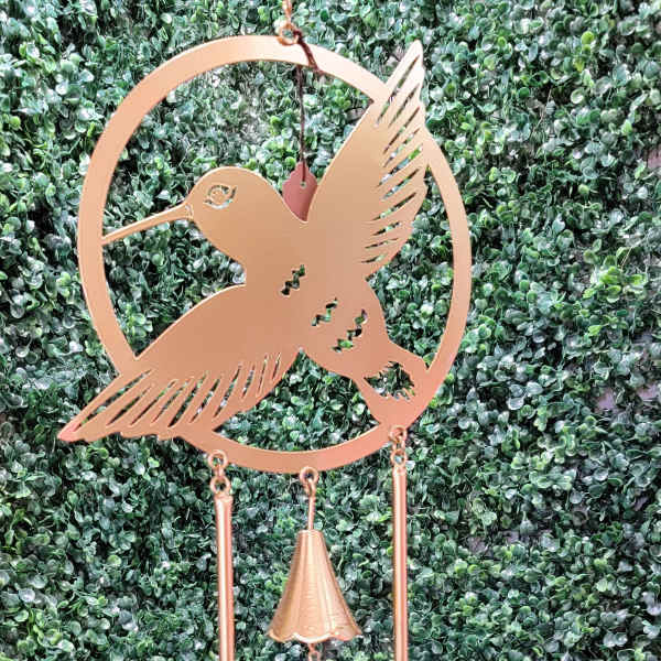 Copper hummingbird wind chime hanging against a green hedge