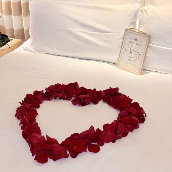 Red rose petals arranged in a heart on a bed with a "Will you marry me?" sign