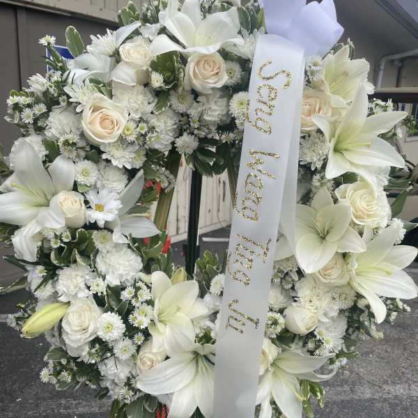 White floral funeral wreath with lilies, roses, and a memorial ribbon