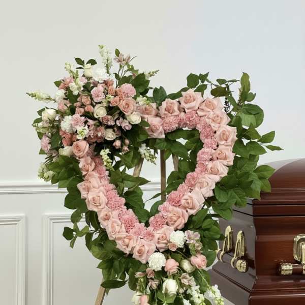 Heart-shaped pink floral funeral wreath on a stand beside a casket