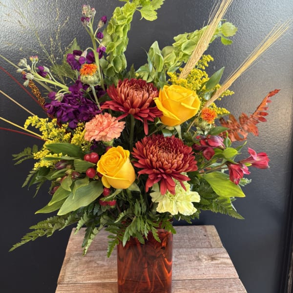 Mixed bouquet with yellow roses, burgundy mums, and purple flowers in an amber vase