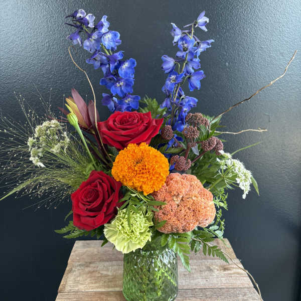 Mixed bouquet with red roses, blue delphinium, and orange blooms in a green glass vase
