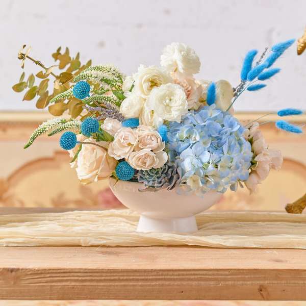 Pastel floral arrangement in a white pedestal bowl with blue accents