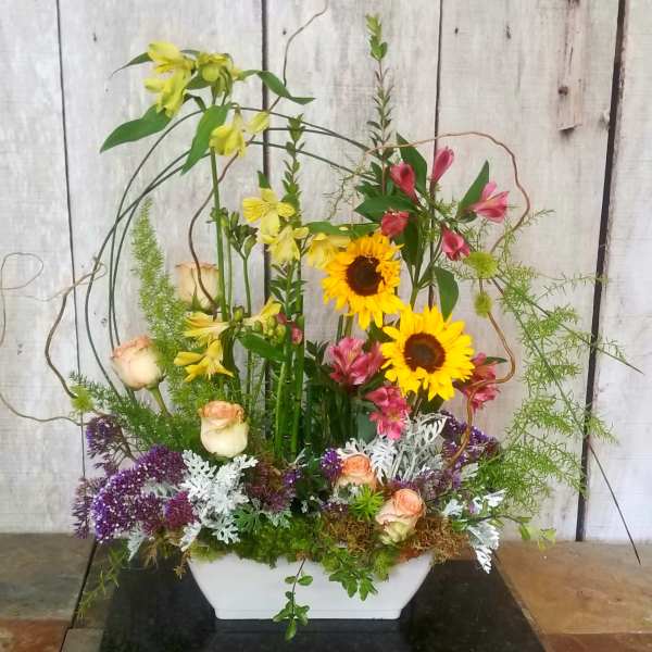 Tall mixed floral arrangement in a white rectangular vase with sunflowers and roses