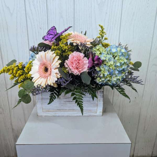 Mixed pastel flowers in a white wooden box with a purple butterfly accent.