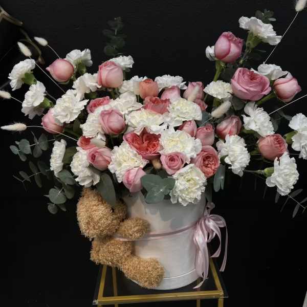 Pink and white roses with carnations in a white hatbox, with a teddy bear attached