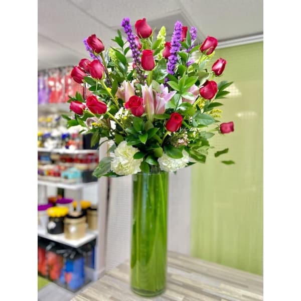 Tall arrangement of red roses, pink lilies, white carnations and purple flowers in a green glass cylinder vase