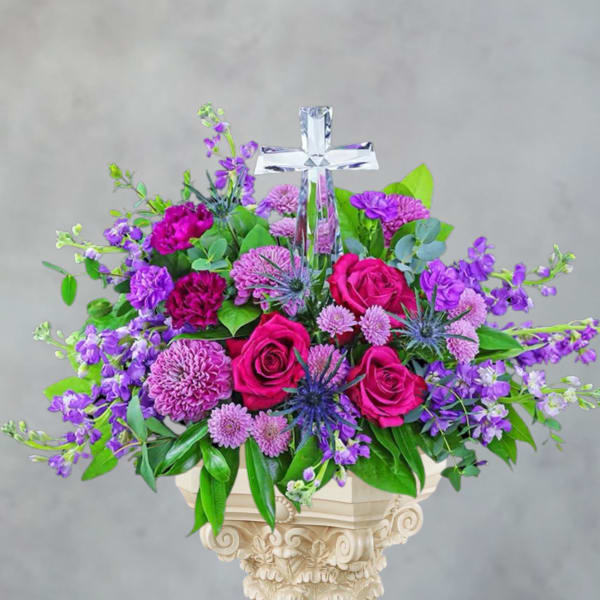 Purple and pink floral arrangement with a clear cross topper in a pedestal vase