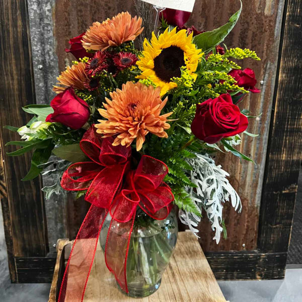 Bouquet of sunflowers, roses, and mums in a glass vase with a red ribbon