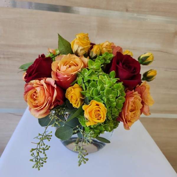 Low glass vase of orange, yellow, and red roses with bright green hydrangea