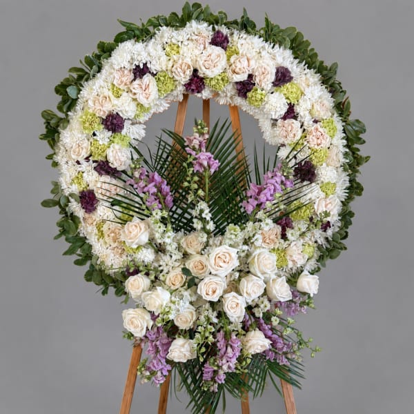 Large floral funeral wreath on a wooden easel with white, cream, purple, and green flowers.