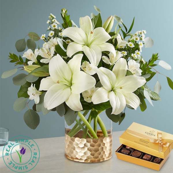 White lilies in a glass vase with a box of chocolates beside it