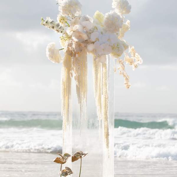 Tall white floral arrangement on a clear pedestal by the beach