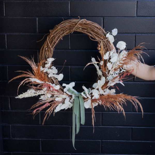 Asymmetrical dried floral wreath with white accents and sage ribbon on a grapevine base.