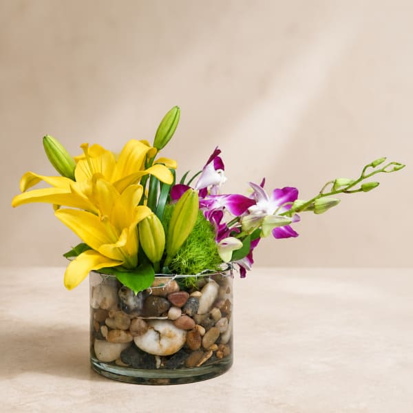 Yellow lilies and purple orchids in a pebble-filled glass vase