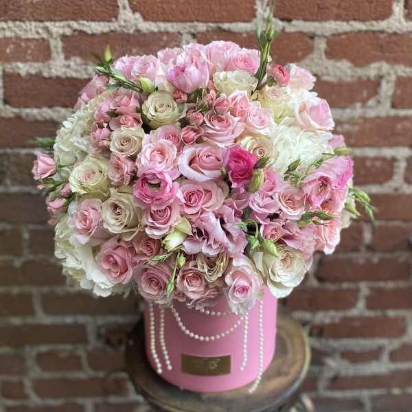 Round arrangement of pale pink and cream roses in a pink hatbox with pearl garlands.