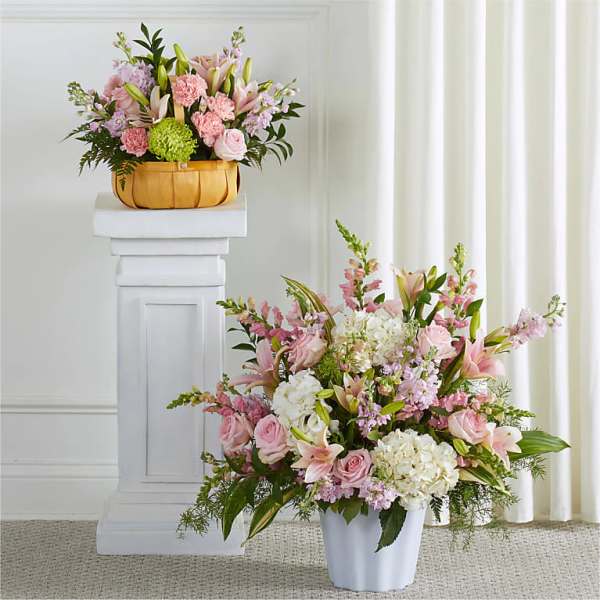 Two pastel floral arrangements, one in a white vase and one in a basket on a pedestal.