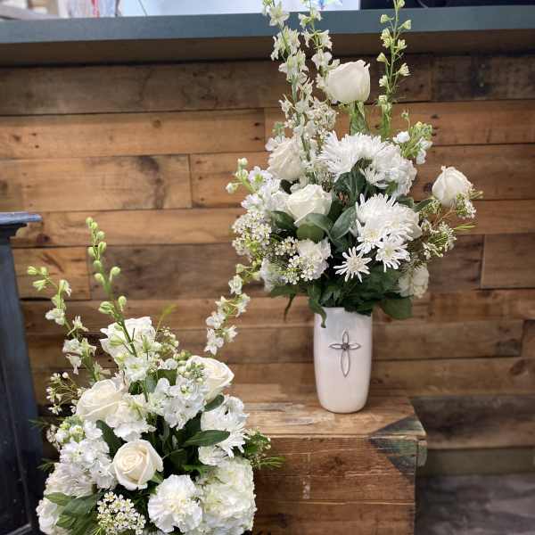 Two white floral arrangements in tall white vases on a wooden surface