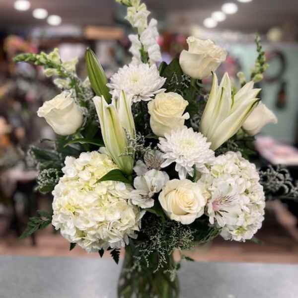 White floral arrangement in a clear glass vase with roses, lilies, and hydrangeas