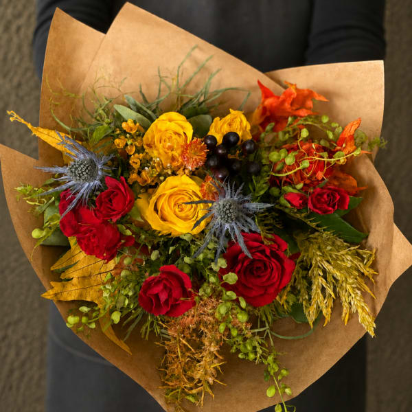 Handheld bouquet of red and yellow roses with blue thistle in brown paper