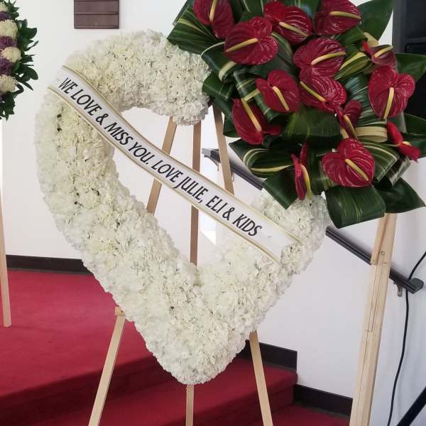 Heart-shaped white floral wreath on an easel with a red anthurium spray.
