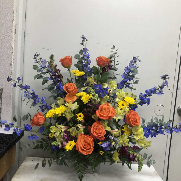 Large mixed bouquet with orange roses, blue flowers, and yellow blooms in a clear vase