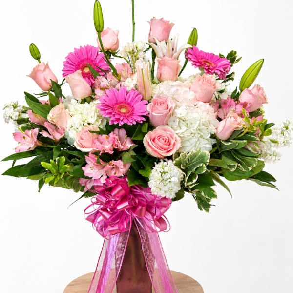 Pink roses, gerbera daisies, lilies, and white hydrangeas in a tall vase with a pink ribbon