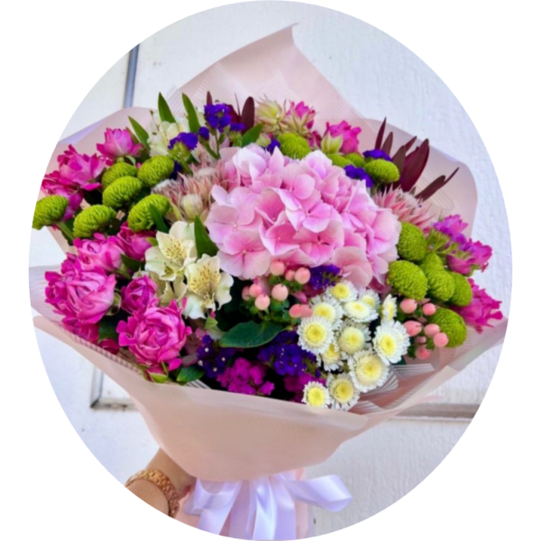 Handheld bouquet of pink, green, white, and purple flowers wrapped in pale paper