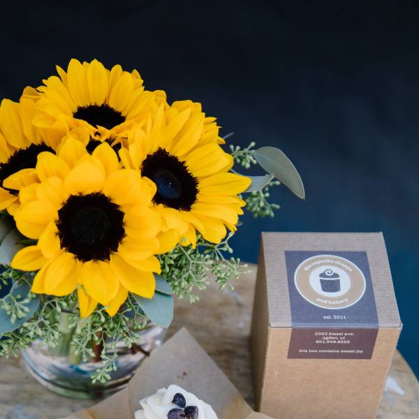Sunflower bouquet in a glass vase beside a cupcake and a bakery box