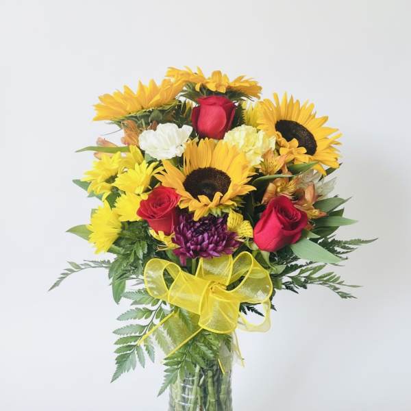 Bouquet of sunflowers, red roses, and mixed blooms in a glass vase with a yellow bow