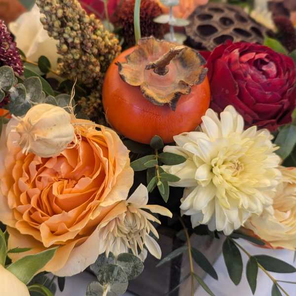 Fall-toned arrangement with orange rose, cream mums, red bloom, and a persimmon in a dark vase.
