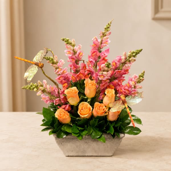 Orange roses and pink snapdragons in a rectangular vase with dragonfly picks