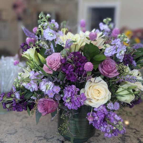 Large arrangement of purple, lavender, and cream flowers, including roses and stock, in a green vase