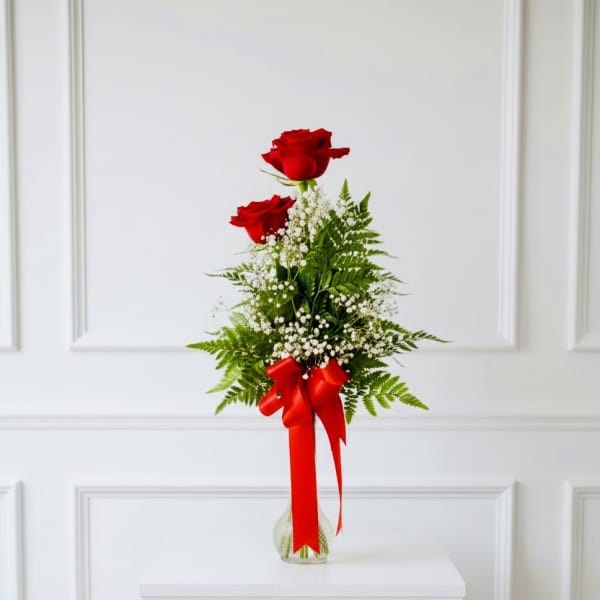 Two red roses in a clear vase with baby's breath and a red ribbon