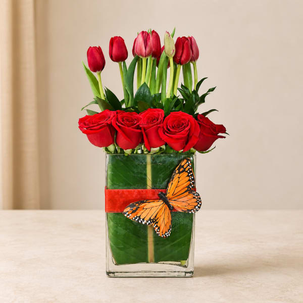 Red roses and tulips arranged in a clear square vase with a butterfly decoration.