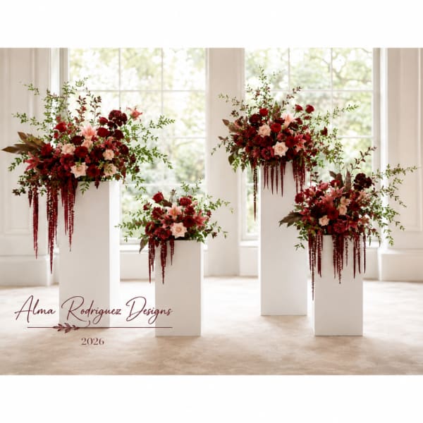 Four red and blush floral arrangements on white pedestals in a bright room