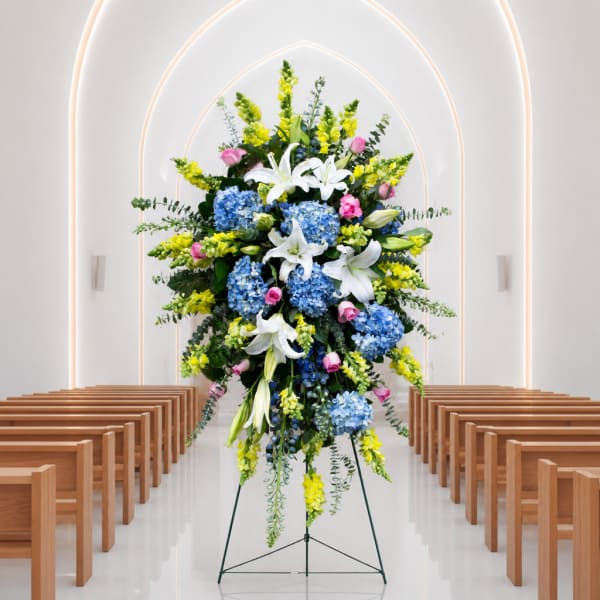 Tall floral spray with white lilies, blue hydrangeas, and yellow blooms on an easel