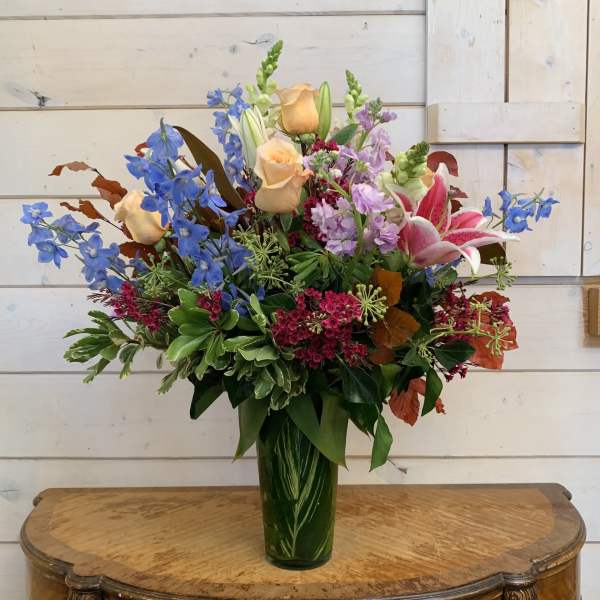 Mixed bouquet of blue delphinium, peach roses, pink lilies, and purple flowers in a green vase