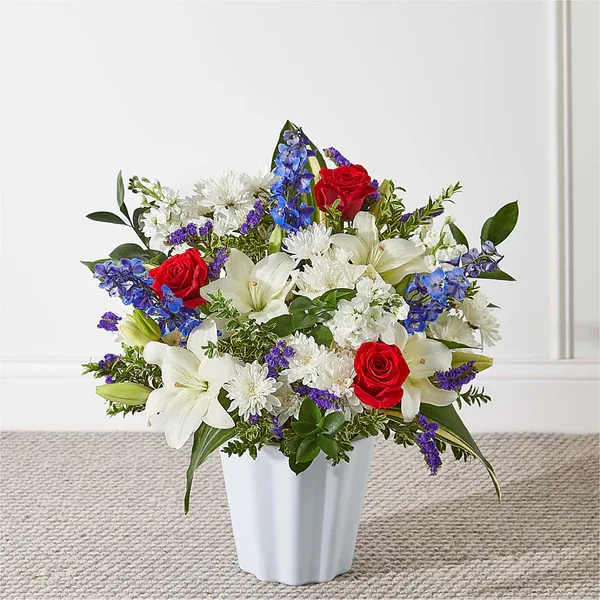 Patriotic-style arrangement of red roses, white lilies and mums, and blue flowers in a white vase