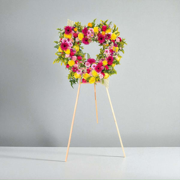 Heart-shaped floral standing wreath on a wooden easel with pink, yellow, and orange blooms