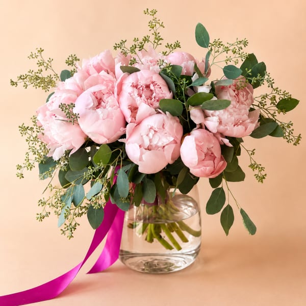 Pink peonies in a clear glass vase with greenery and a magenta ribbon