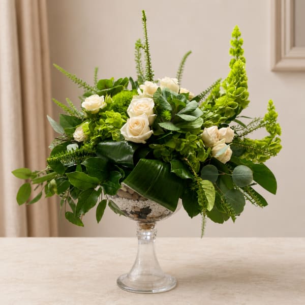 White roses in a clear pedestal vase with lush green foliage
