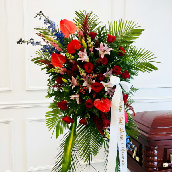 Tall funeral spray with red roses, lilies, and tropical leaves beside a casket