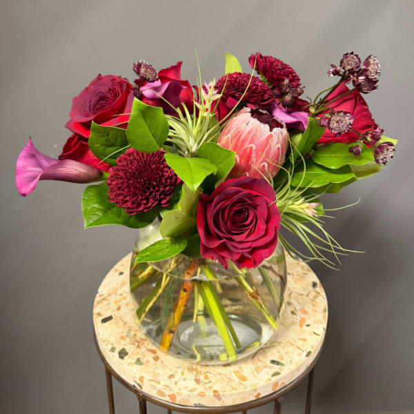 Bouquet of red roses, burgundy blooms, and a pink protea in a glass vase
