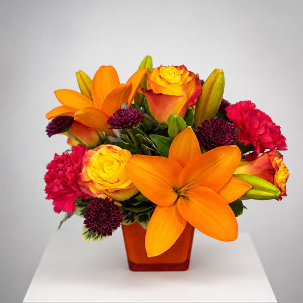 Orange lilies, yellow roses, and pink carnations in an amber vase