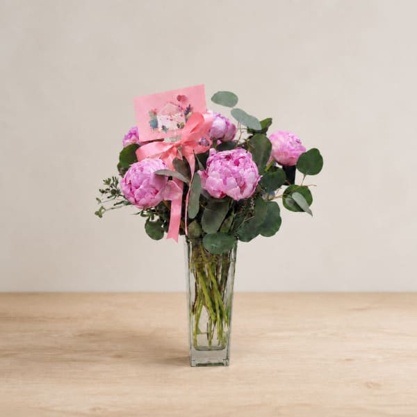 Pink peony bouquet in a clear glass vase with a ribbon and card