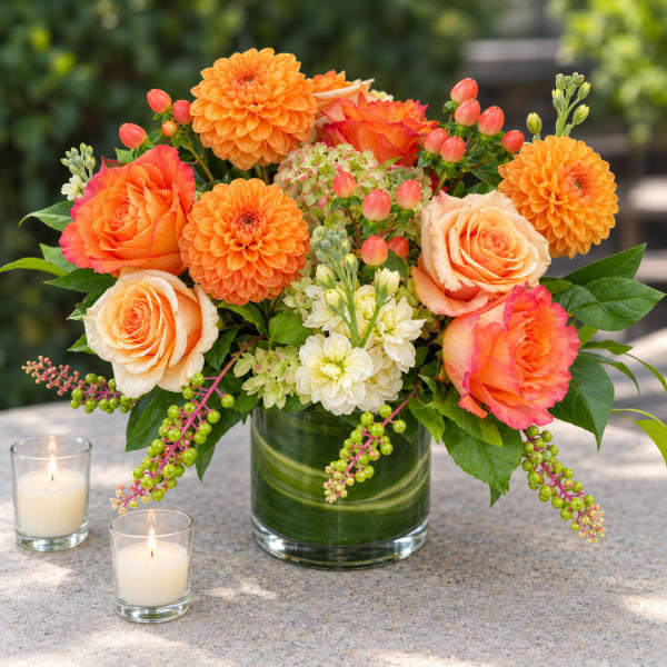 Orange roses and dahlias arranged in a glass vase with candles nearby