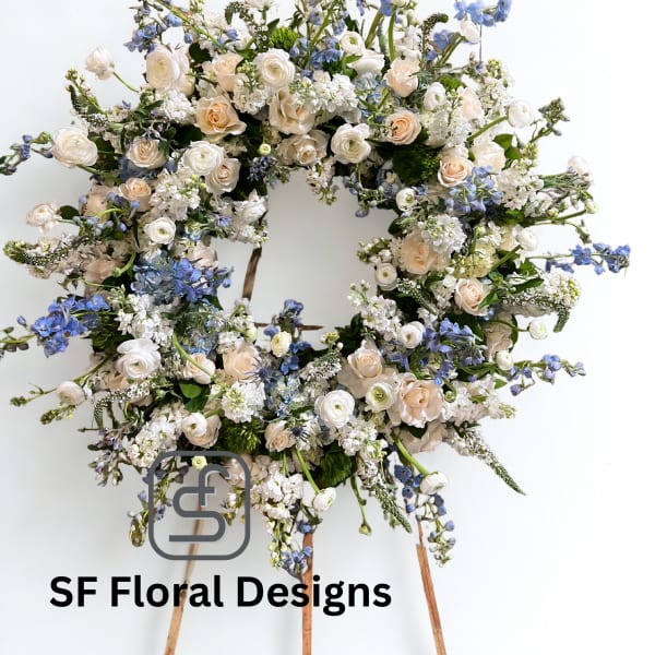 Large round wreath of cream roses and white flowers with soft blue accents on a wooden easel.