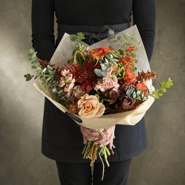 Handheld bouquet of peach, red, and pink flowers wrapped in paper